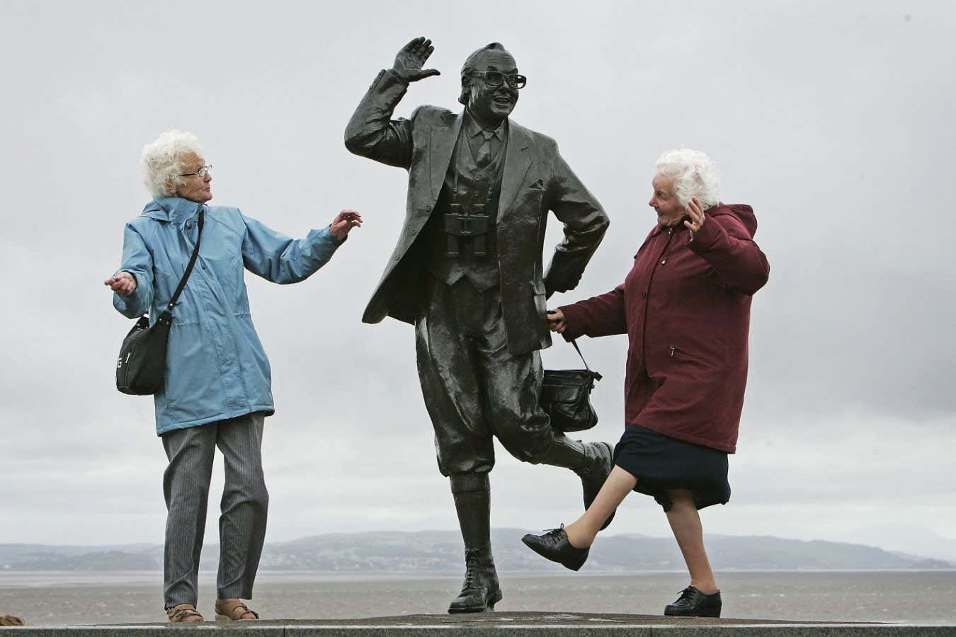 MORCAMBE, UNITED KINGDOM - JUNE 22: Despite inclement weather pensioners raise a happy smile as they perform the famously British dance of comedians Morcambe and Wise next to a statue of Eric Morcambe, at Morcambe Bayon June 22, 2006, in Morcambe, England. Confidence & Happiness specialist, Scientist Cliff Arnall from the University of Cardiff has identified June 23, 2006 as being the happiest day of the year. His calculations were based on outdoor activity, nature, social interaction, childhood summers, positive memories, temperature and holidays. (Photo by Christopher Furlong/Getty Images)