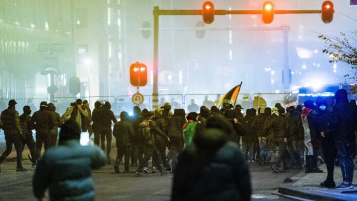 Protesters, many wearing dark clothing and masks, gather in the street during a pro-Palestinian demonstration in Amsterdam. Some carry flags, with a backdrop of urban lighting and visible police presence